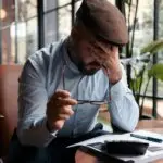 A man sitting at a table, holding his head in stress, facing tax papers and a calculator.