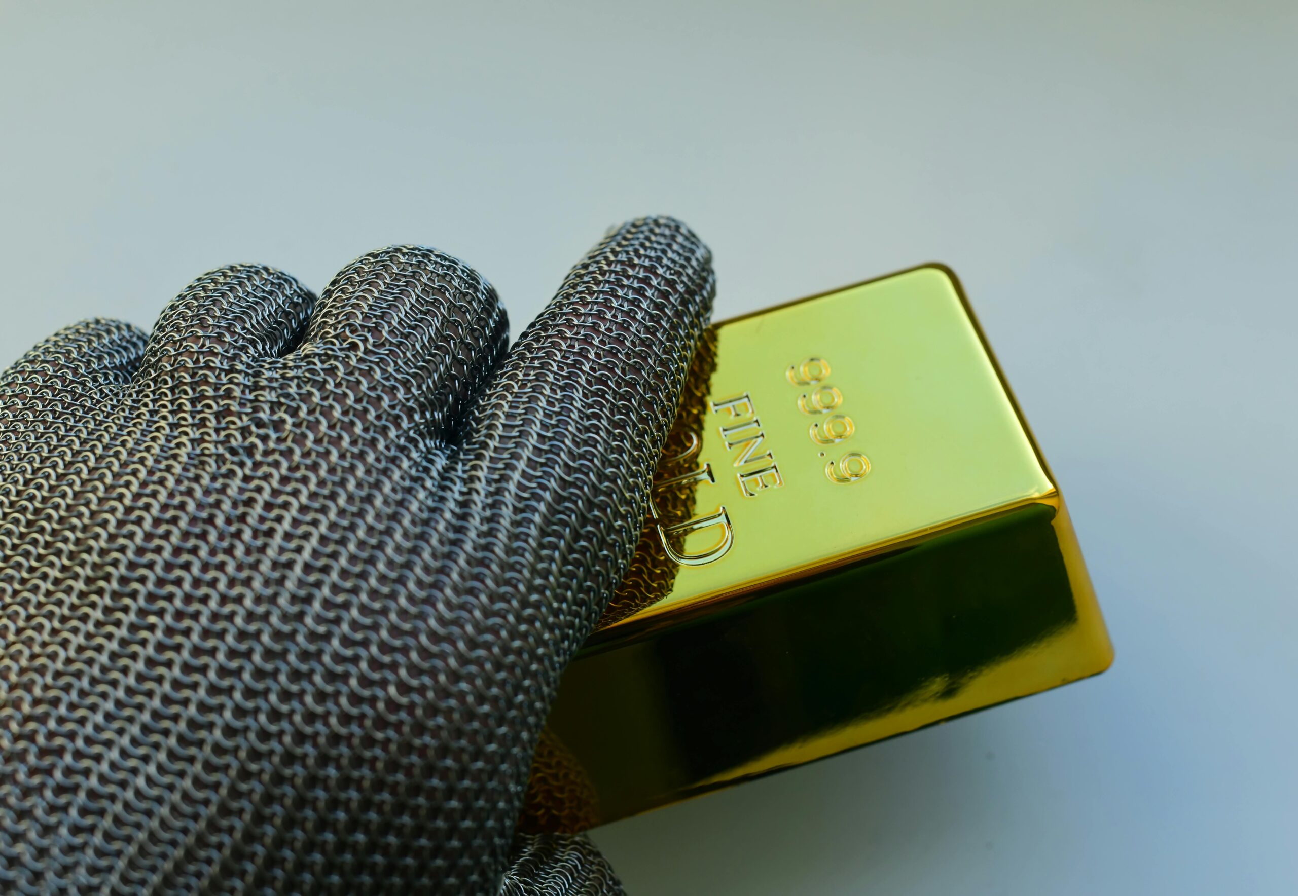 Close-up of a metal gloved hand holding a shiny gold bar indoors.